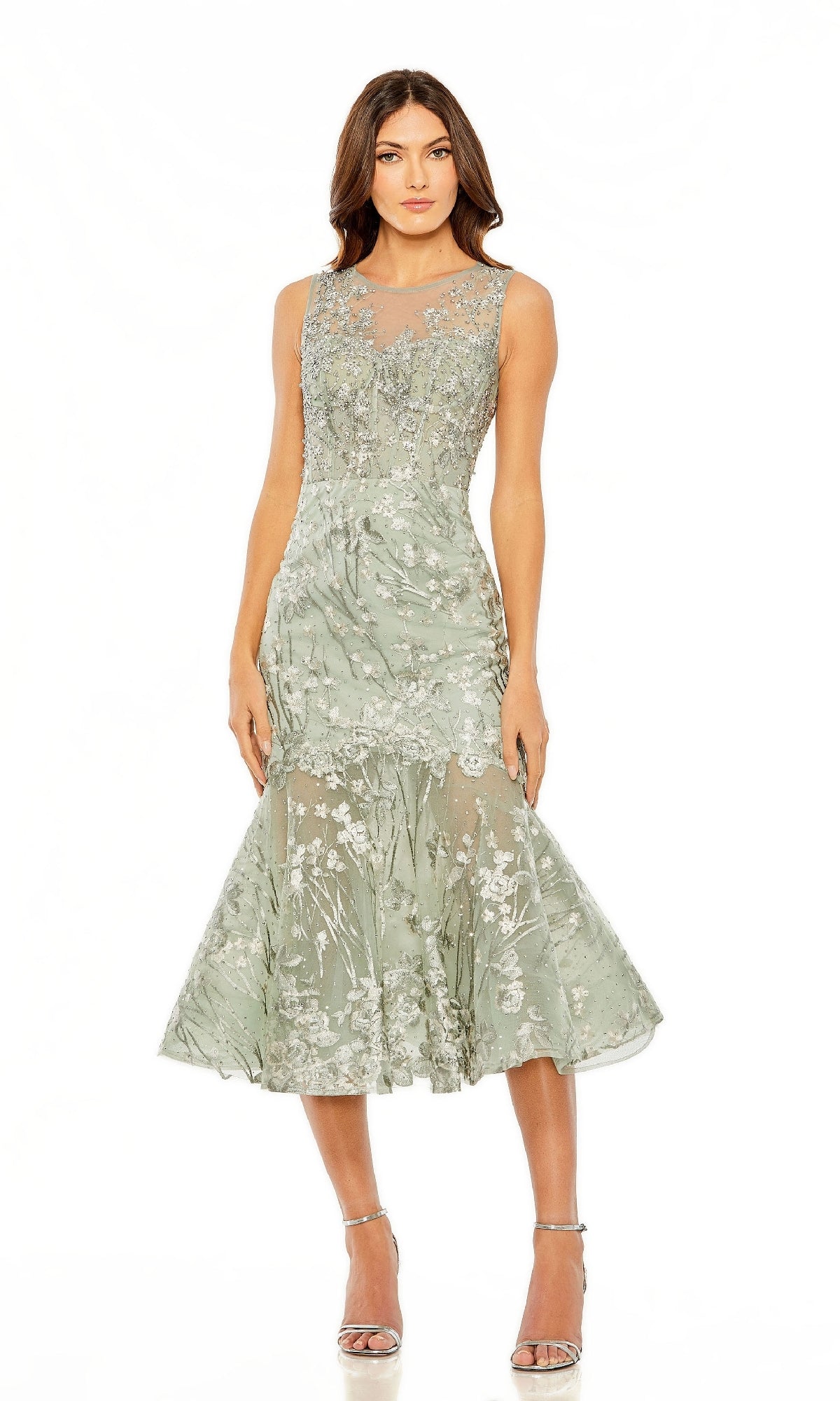 A woman poses in the Sage Midi Wedding Guest Dress: Mac Duggal 20646—a sleeveless, sage green floral lace midi with sheer details, drop-waist silhouette, flared hem, and silver high heel sandals.